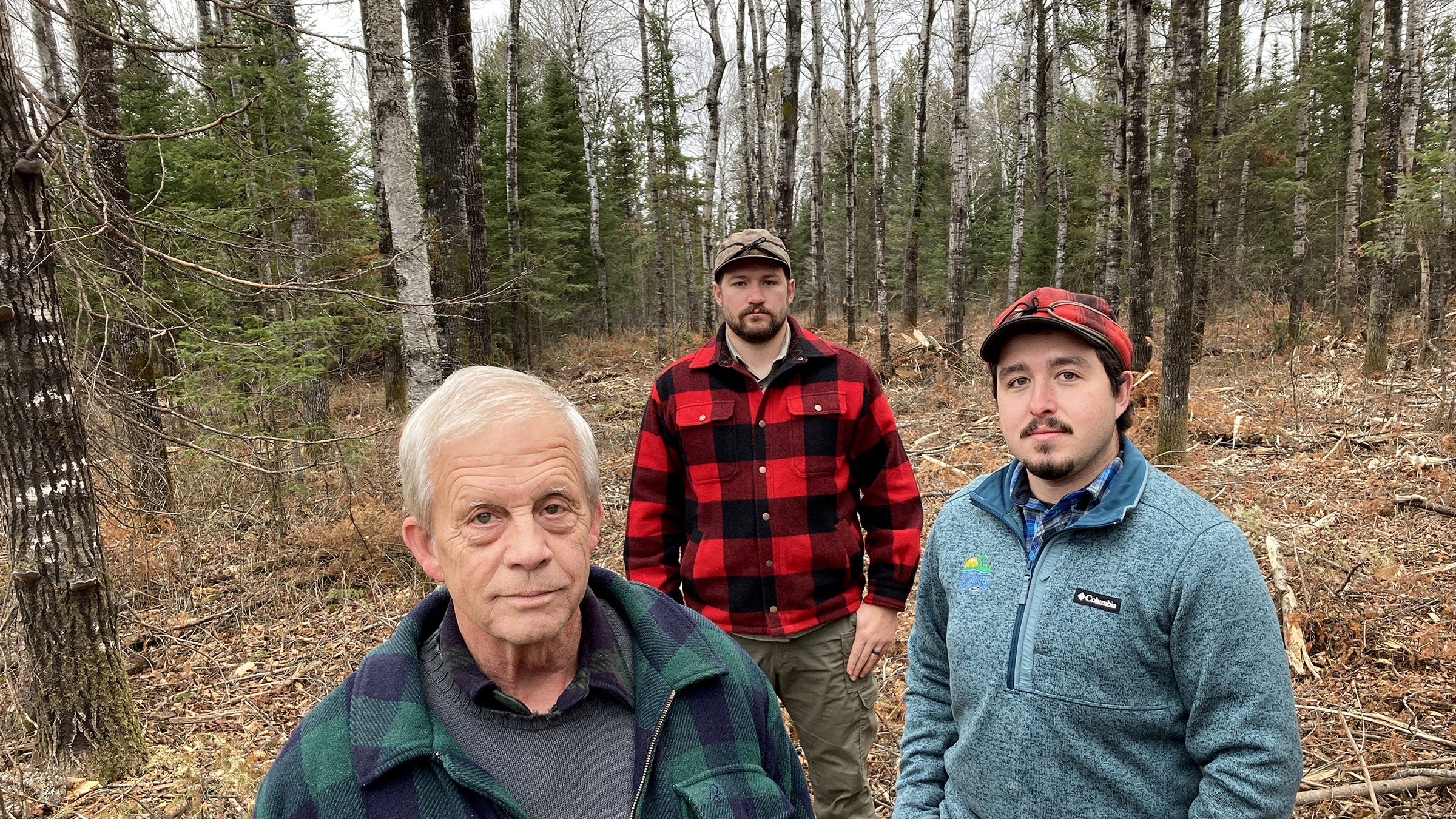 Three men stand in a clearing in the woods