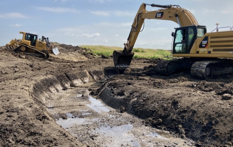 A backhoe excavates a stream channel. A bulldozer is in the background.